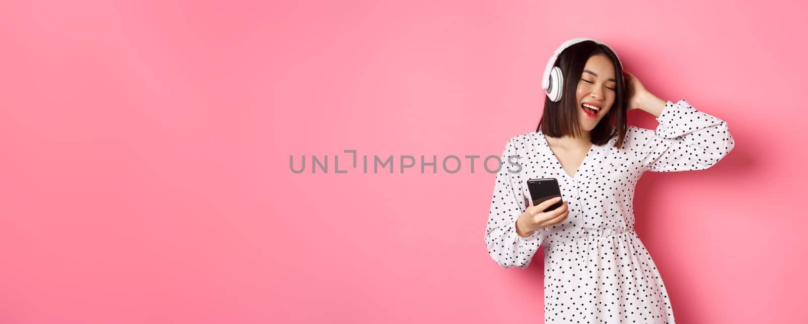 Carefree asian woman having fun, dancing and listening music in headphones, holding mobile phone, standing in dress over pink background.