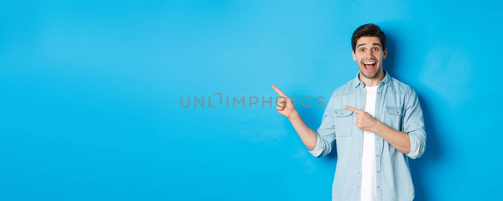 Excited handsome 25s years man with beard, pointing fingers left and smiling amazed, standing against blue background.
