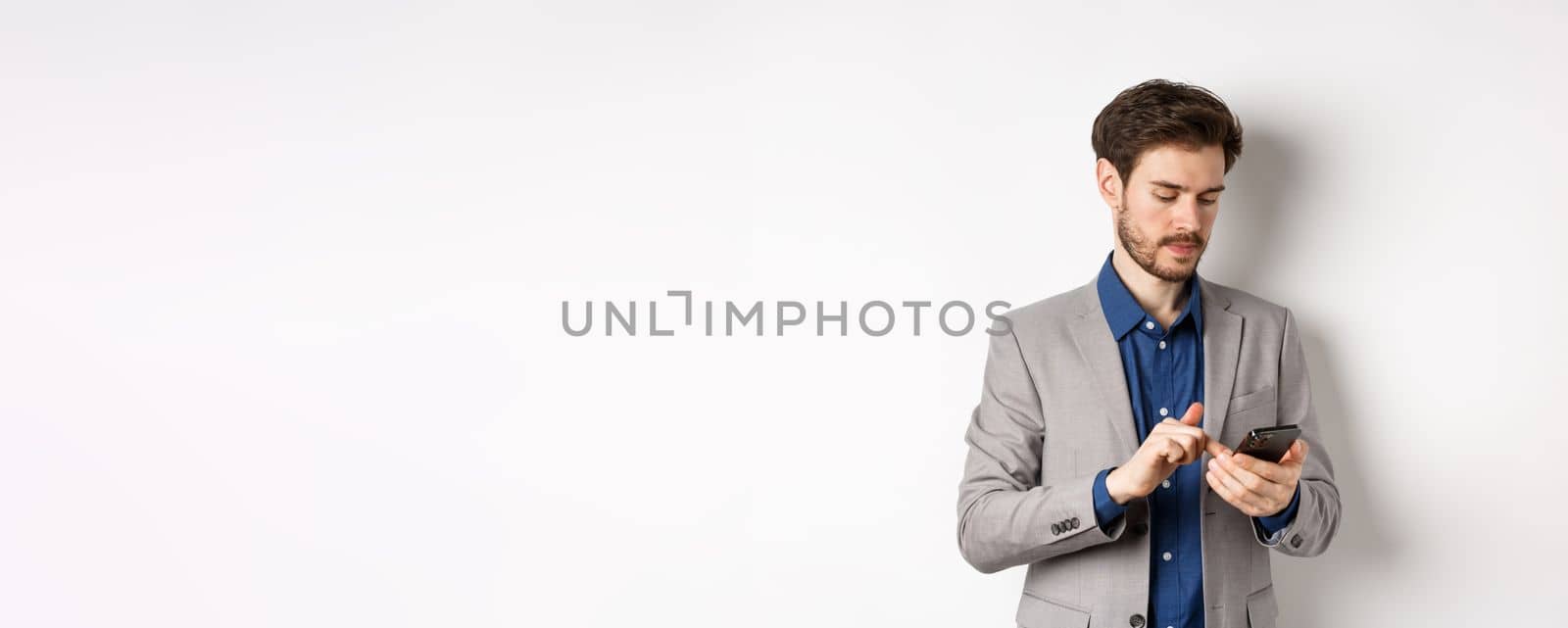 Handsome businessman in stylish suit chatting on smartphone, using mobile phone, standing on white background.