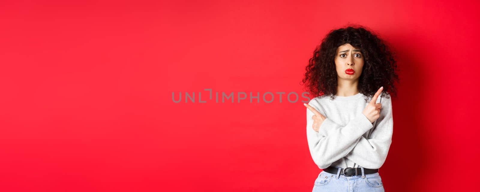 Nervous sad girl with curly hair, sobbing miserable and pointing fingers sideways, showing two things and look indecisive, red background.