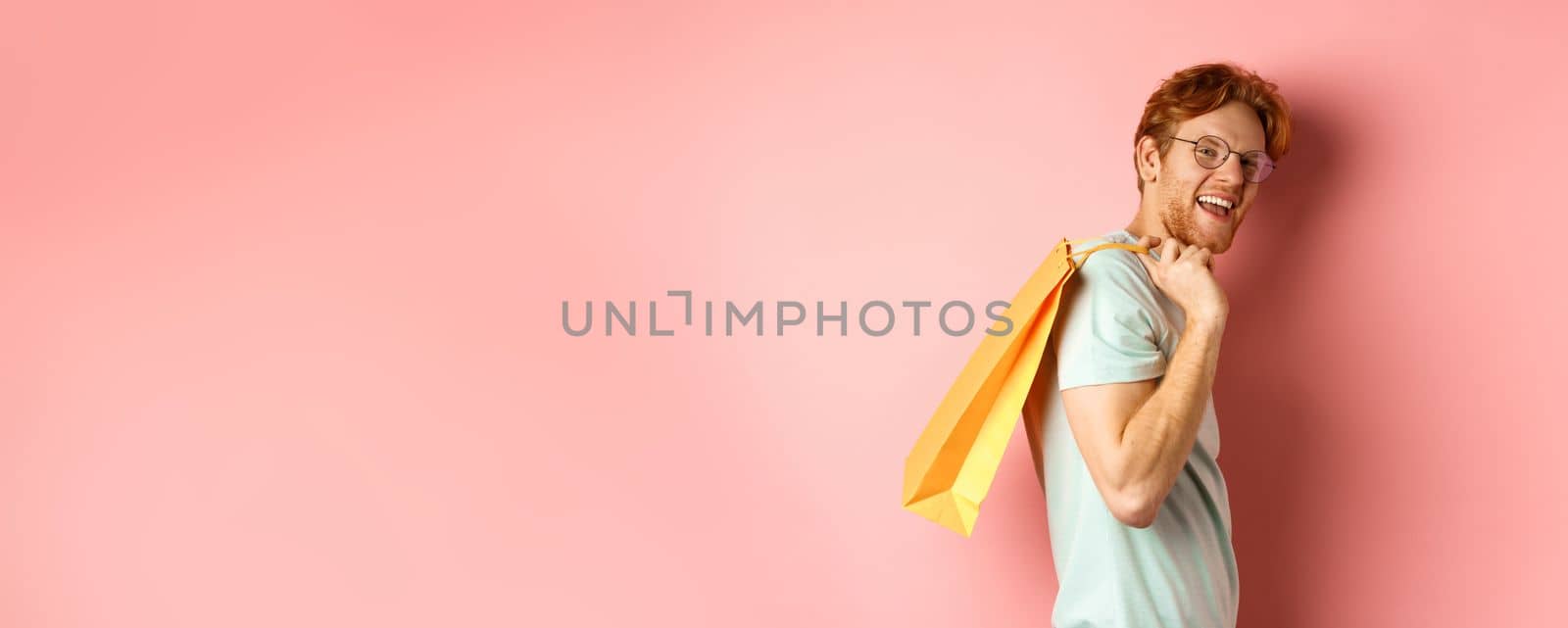 Carefree young man with red hair and glasses, walking with shopping bag over his shoulder and smiling, shopper buying presents, standing satisfied over pink background.