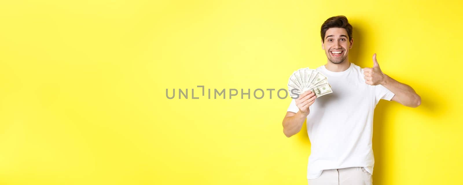 Satisfied buyer man showing thumb-up and holding money, shopping with cash, standing over yellow background by Benzoix