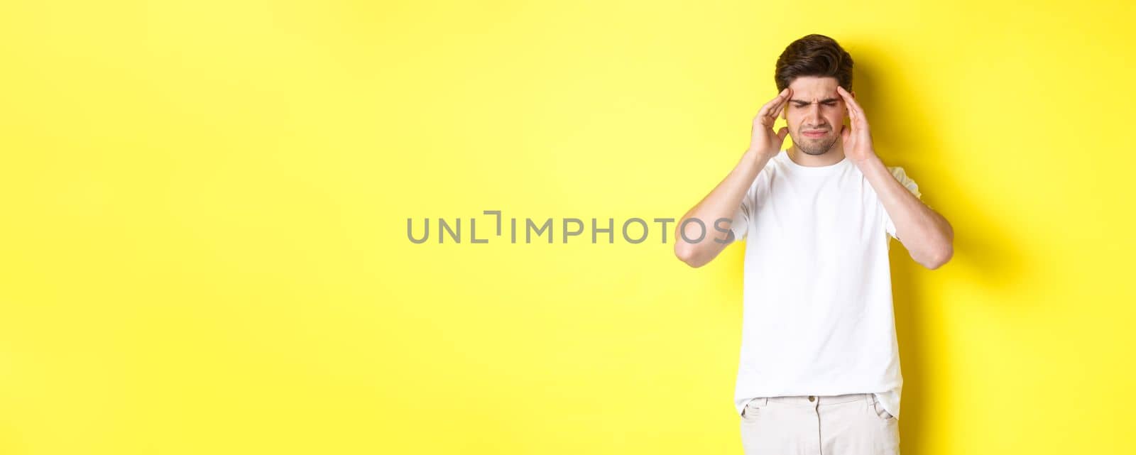 Troubled man touching head and grimacing from pain, complaining on headache, standing over yellow background. Copy space