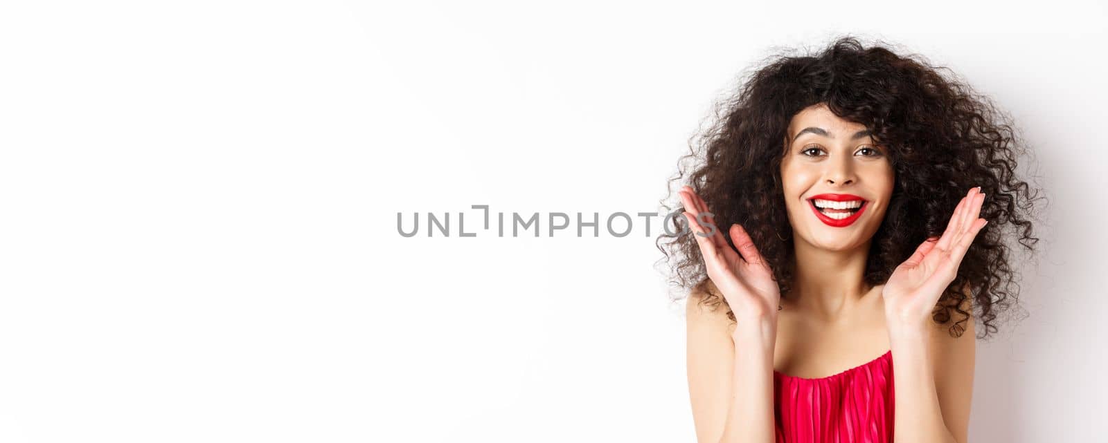 Close-up of fashionable woman with curly hair and red dress, clap hands smiling happy, applause you, standing on white background.