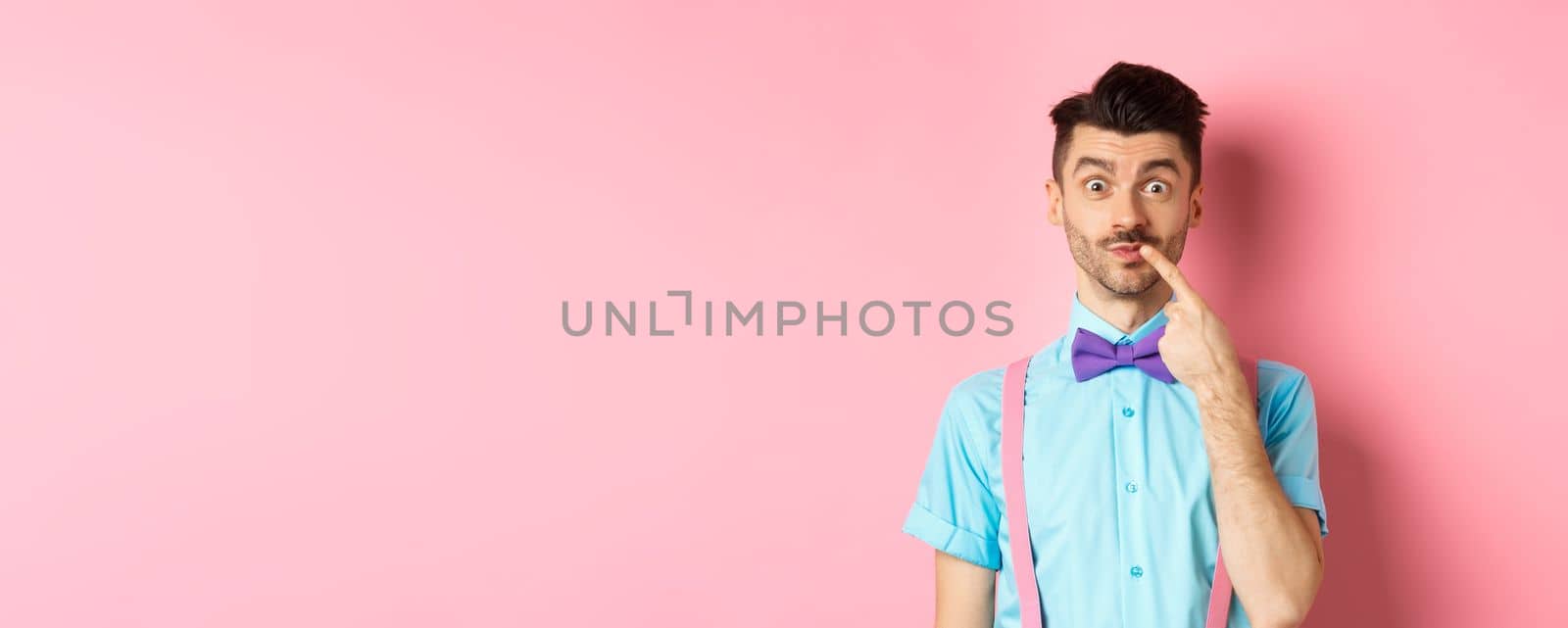 Intrigued happy guy touching lip, looking at camera pensive, making choice what to buy, standing over pink background by Benzoix