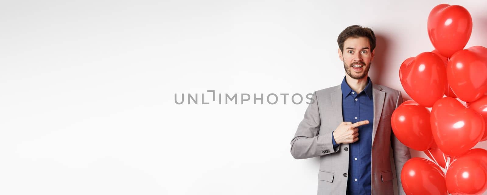 Lovers day. Excited male model in suit pointing finger at Valentines heart balloons and smiling, prepare romantic gifts on date, standing over white background.