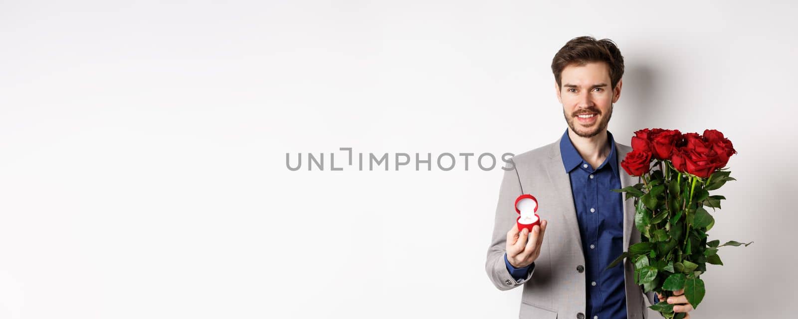 Smiling boyfriend making a marriage proposal, standing with engagement ring and bouquet of red roses, going on romantic Valentines day date, white background.