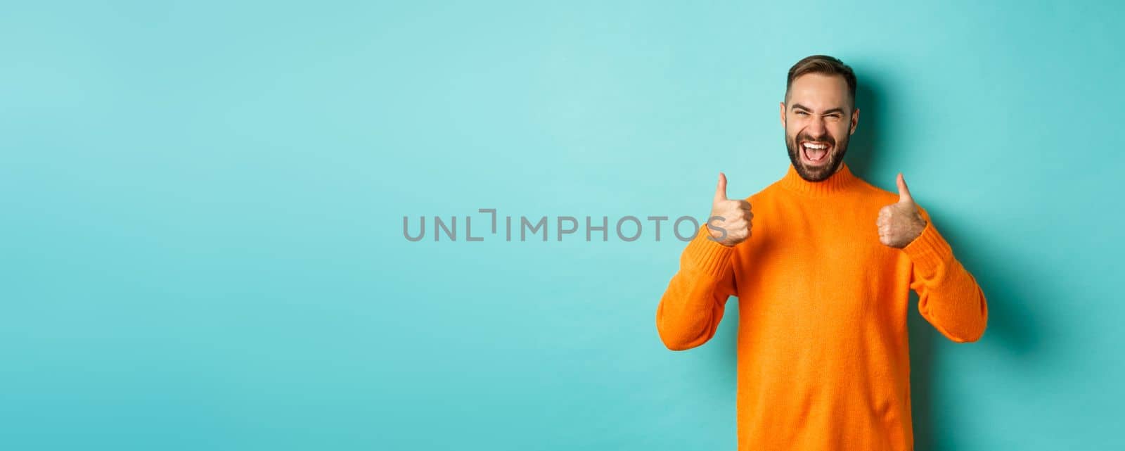 Super good. Satisfied happy man showing thumbs up, agree with you, praise excellent work, looking pleased, standing in orange sweater against light blue background.