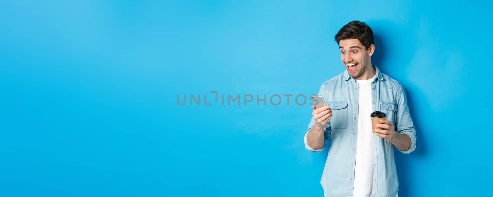 Man looking surprised at smartphone, drinking coffee, standing over blue background amazed by Benzoix