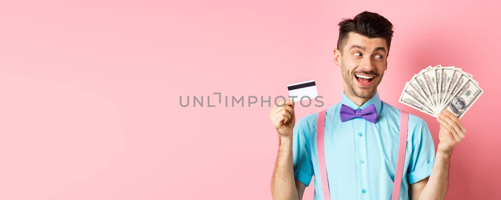 Image of smiling young man in bow-tie holding plastic credit card and looking happy at money, choosing cash, standing over pink background.