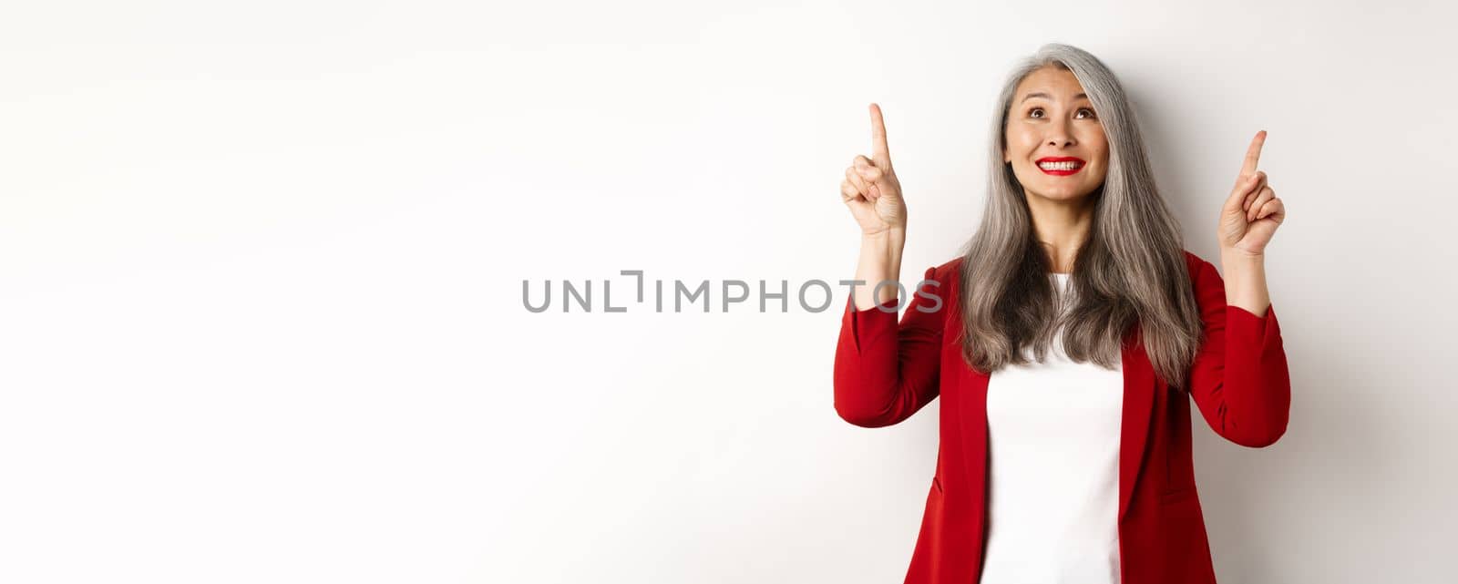 Happy asian lady in red blazer and makeup, looking and pointing fingers up, showing special offer, standing over white background by Benzoix