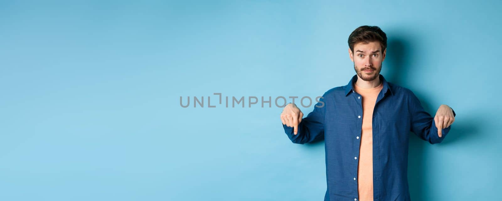 Confused male model in casual clothes, looking puzzled and pointing fingers down, showing banner, standing on blue background.