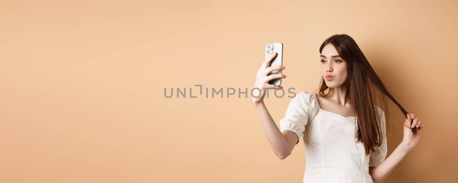 Stylish young woman playing with hair and taking selfie on smartphone, make photo for social media, standing on beige background.