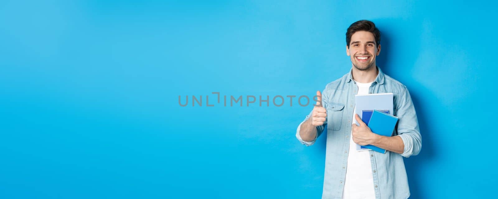 Young man student with notebooks, showing thumb up in approval, smiling satisfied, blue studio background.