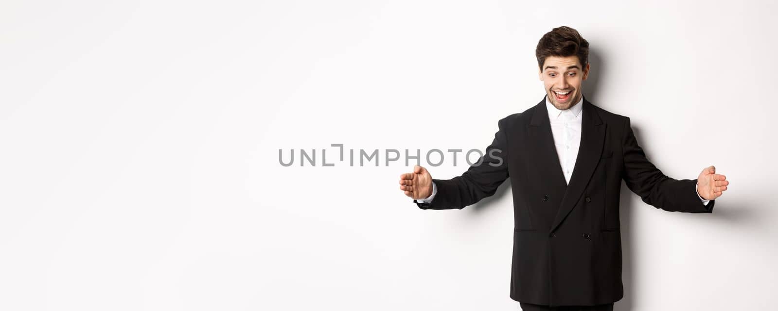 Portrait of excited handsome man in suit, shaping big object on copy space and smiling amazed, holding something, standing over white background by Benzoix