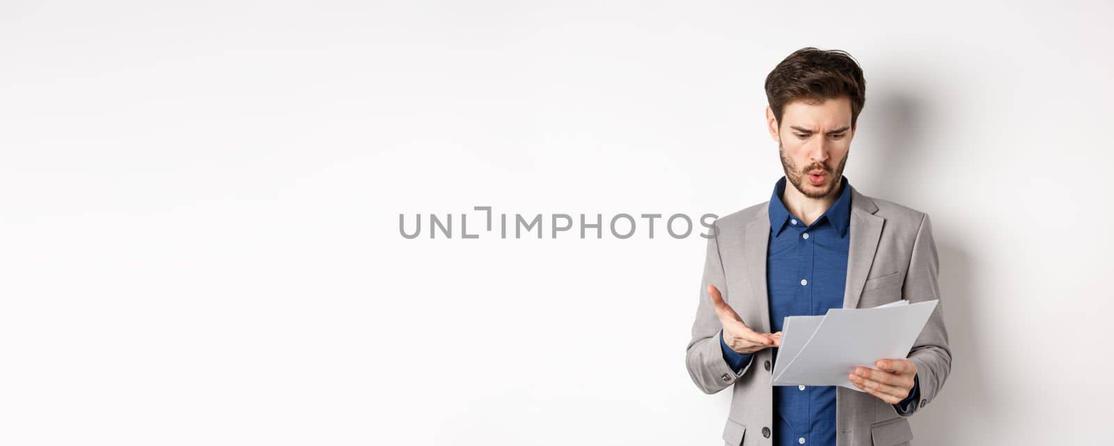 Confused businessman looking at bad document, complaining on deal, pointing at paper frustrated, standing in suit on white background.