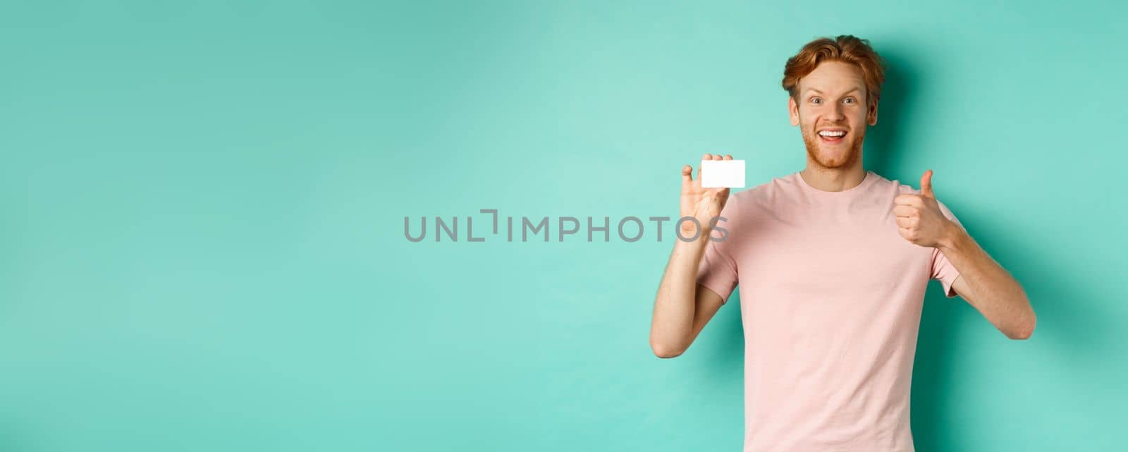 Cheerful male bank client in t-shirt showing thumb up and plastic credit card, smiling satisfied at camera, standing over turquoise background.