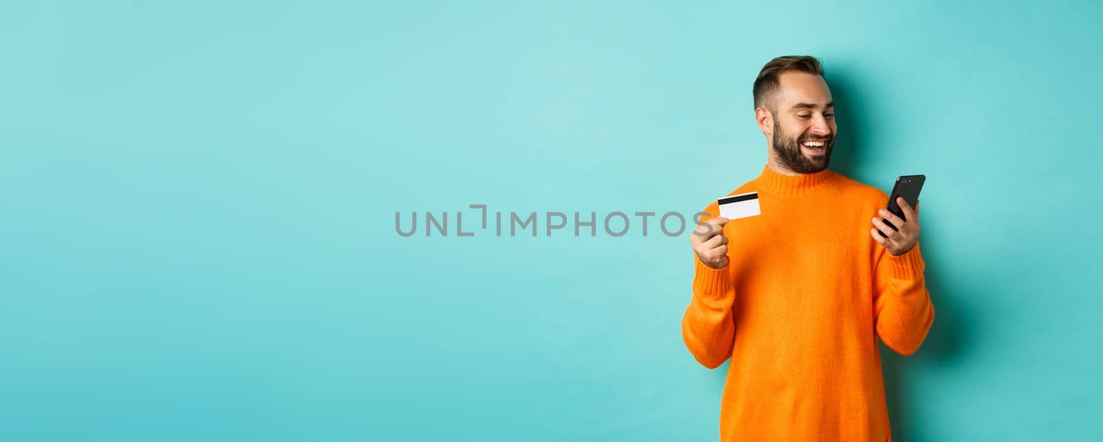 Online shopping. Handsome bearded man paying in internet, holding credit card and stare at mobile screen, standing over turquoise background by Benzoix