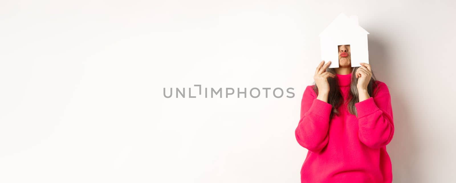 Real estate. Funny asian middle-aged woman hiding face behind paper house model and showing puckered lips, having fun, standing over white background.