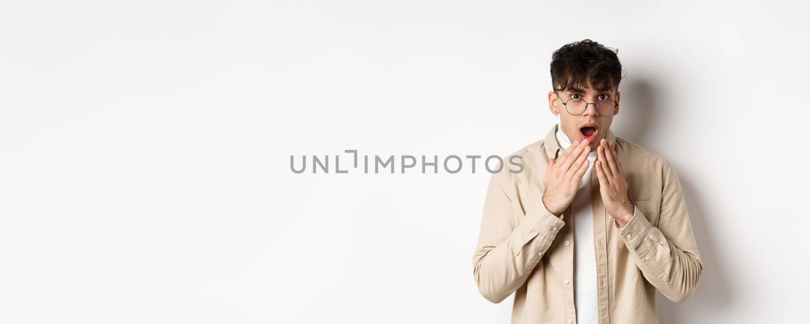 Portrait of handsome guy student looking shocked, hear gossip and stare with disbelief, covering opened mouth with hands, standing on white background.