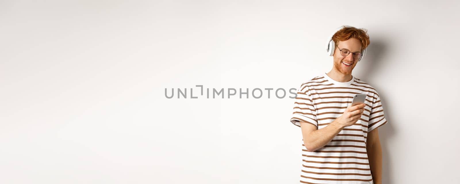 Technology concept. Young redhead hipster in glasses, listening music in headphoes, reading message on smartphone, white background.