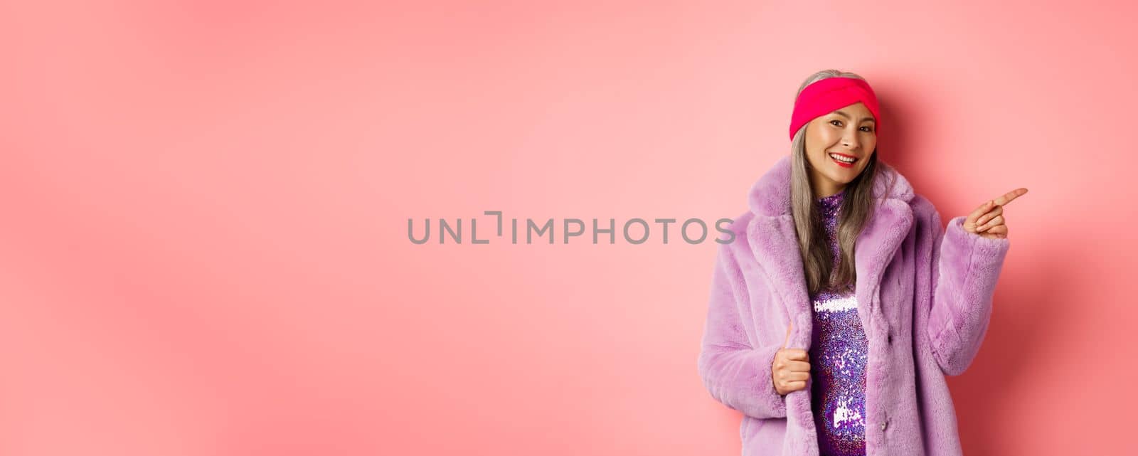 Smiling senior woman looking trendy in winter fur coat, pointing finger right and showing advertisement, standing against pink background.