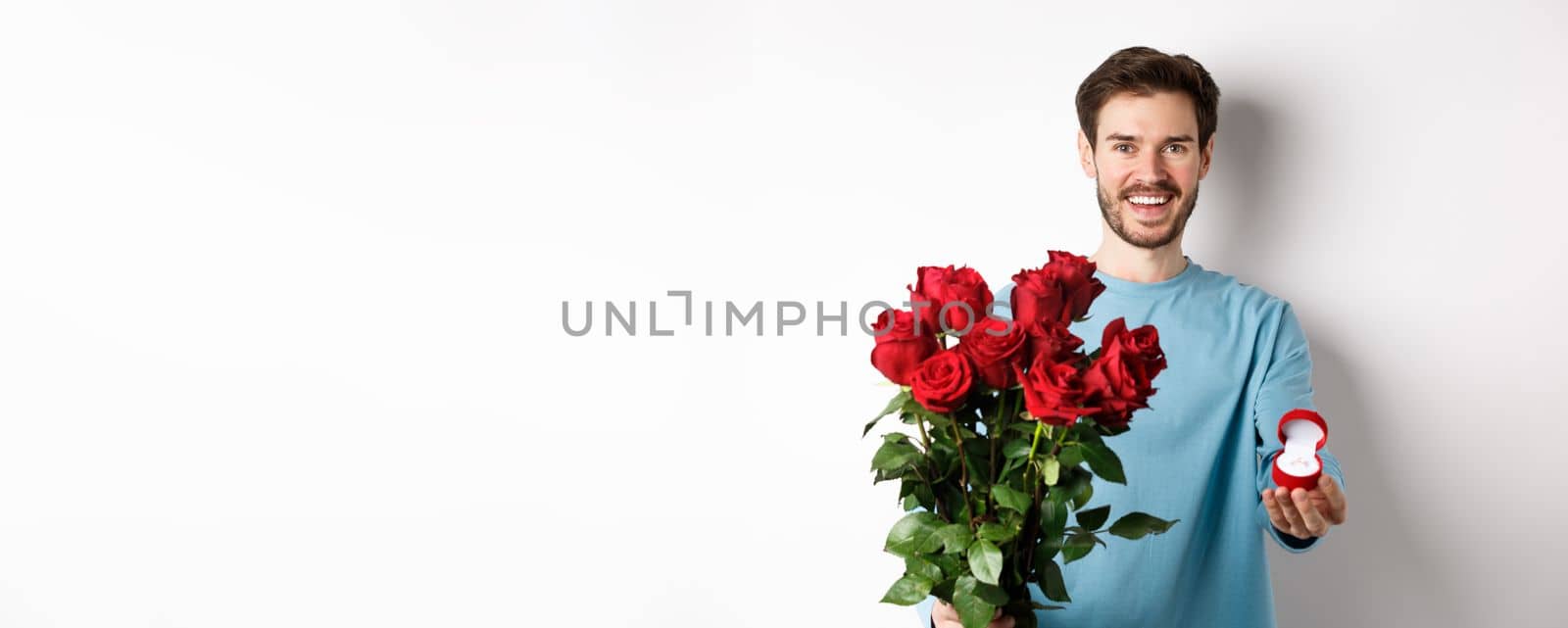 Handsome young man boyfriend making a proposal on Valentines lovers day, holding bouquet of red roses and engagement ring, concept of wedding and relationship.