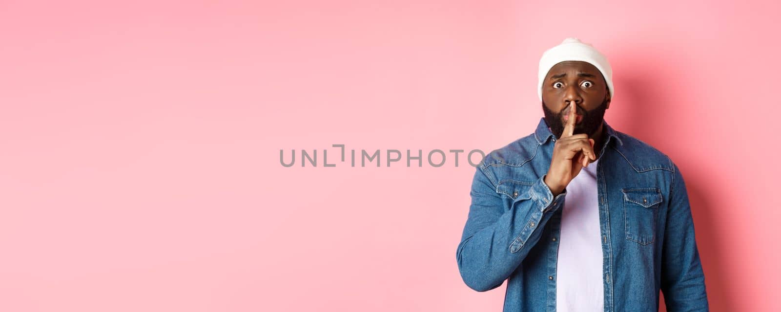Worried Black man asking to keep quiet, sharing secret and hushing at you, holding finger pressed to lips and staring nervously at camera, pink background by Benzoix