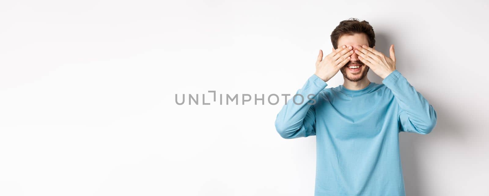 Smiling caucasian man waiting for surprise with closed eyes, expect gift, standing over white background. Celebration and holidays concept.