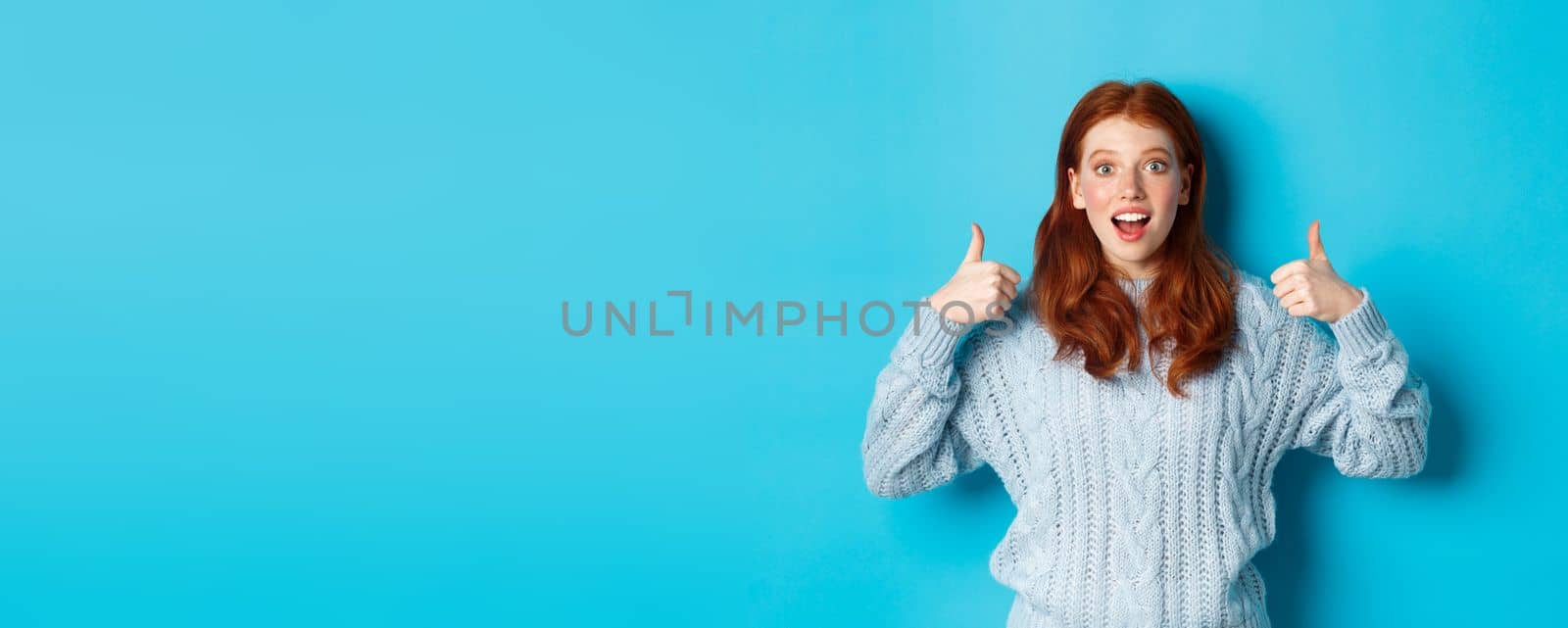 Amazed redhead girl in sweater, showing thumbs up and praising product, looking at camera amazed, standing over blue background. Copy space