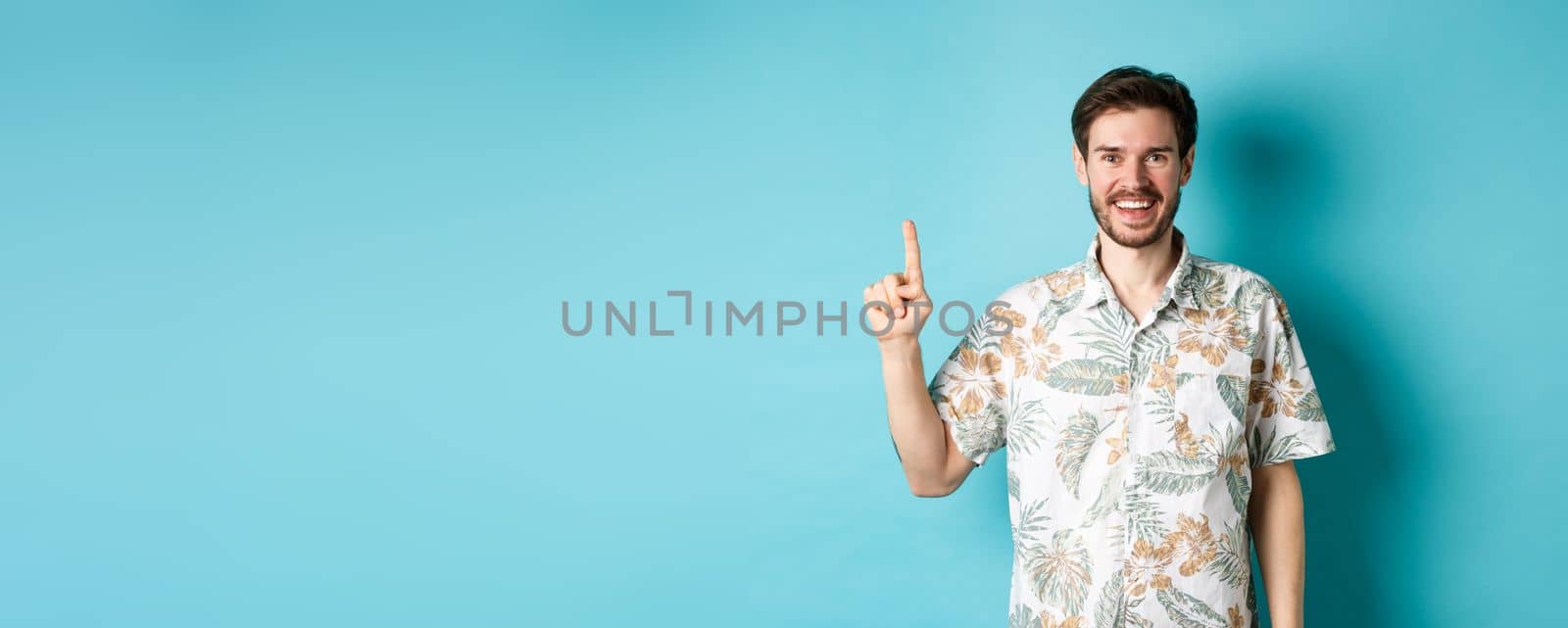 Happy tourist in hawaiian shirt smiling and pointing finger up, showing logo promotion, standing on blue background.