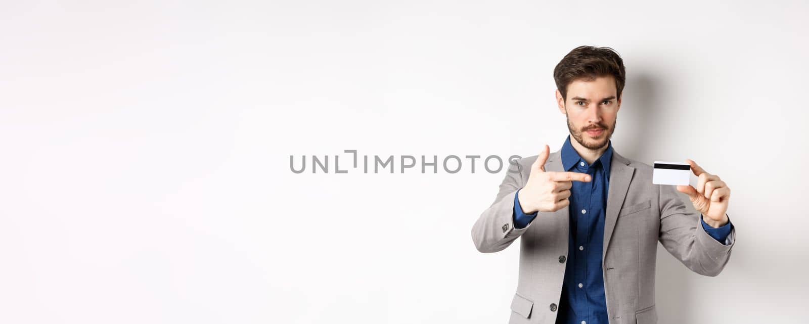 Successful businessman in suit pointing at plastic credit card and smiling, recommending bank, standing on white background.