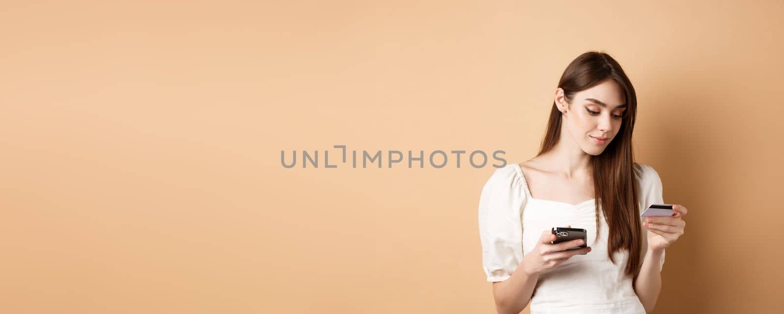 Smiling young woman paying online, looking at credit card and making purchase on mobile phone, shopping in internet, standing on beige background.