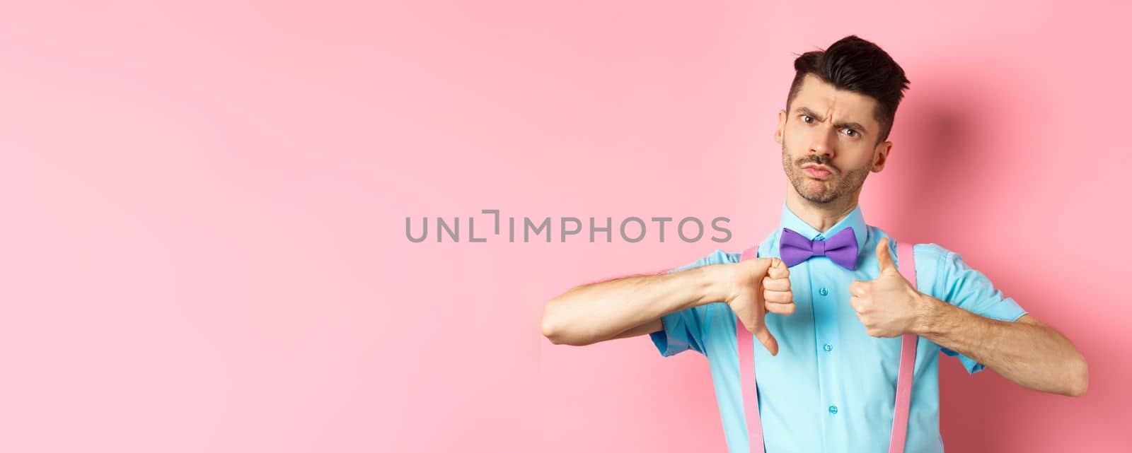 Skeptical young man in bow-tie grimacing, showing thumbs up and down, standing indecisive and judging something, standing over pink background.