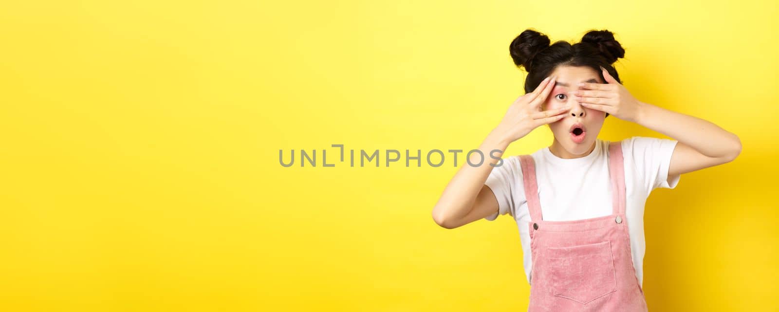 Excited asian teen girl covering eyes with hands and peeking through fingers, gasping amazed at camera, standing in summer clothes on yellow background by Benzoix