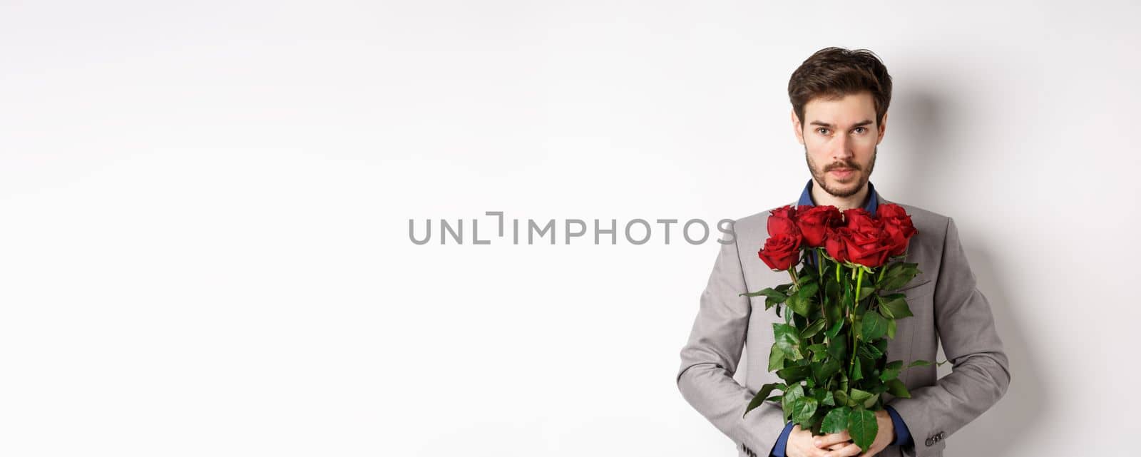 Valentines day and love concept. Confident and handsome man in suit holding bouquet of roses, looking at camera, standing over white background by Benzoix
