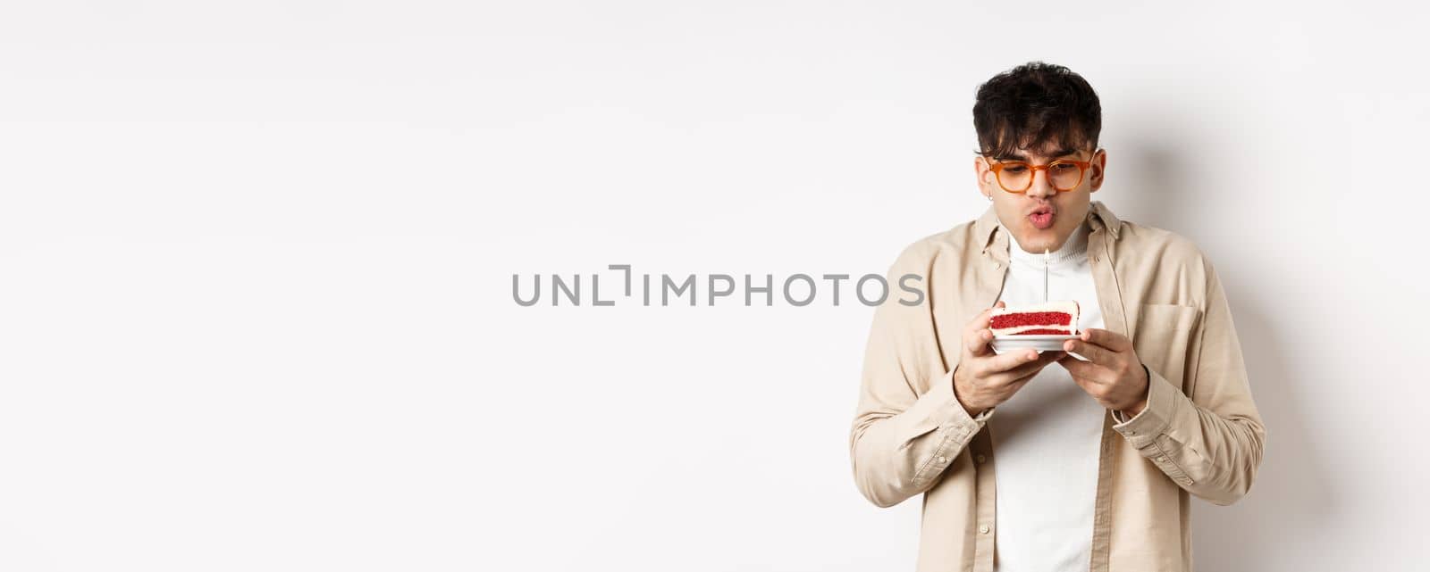 Funny young guy in glasses blowing candle and making wish on birthday cake, standing cheerful against white background.