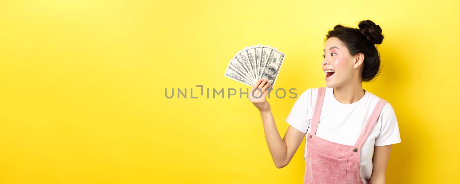 Shopping. Rich and stylish asian female model showing money, looking at dollar bills with happy face, standing on yellow background.