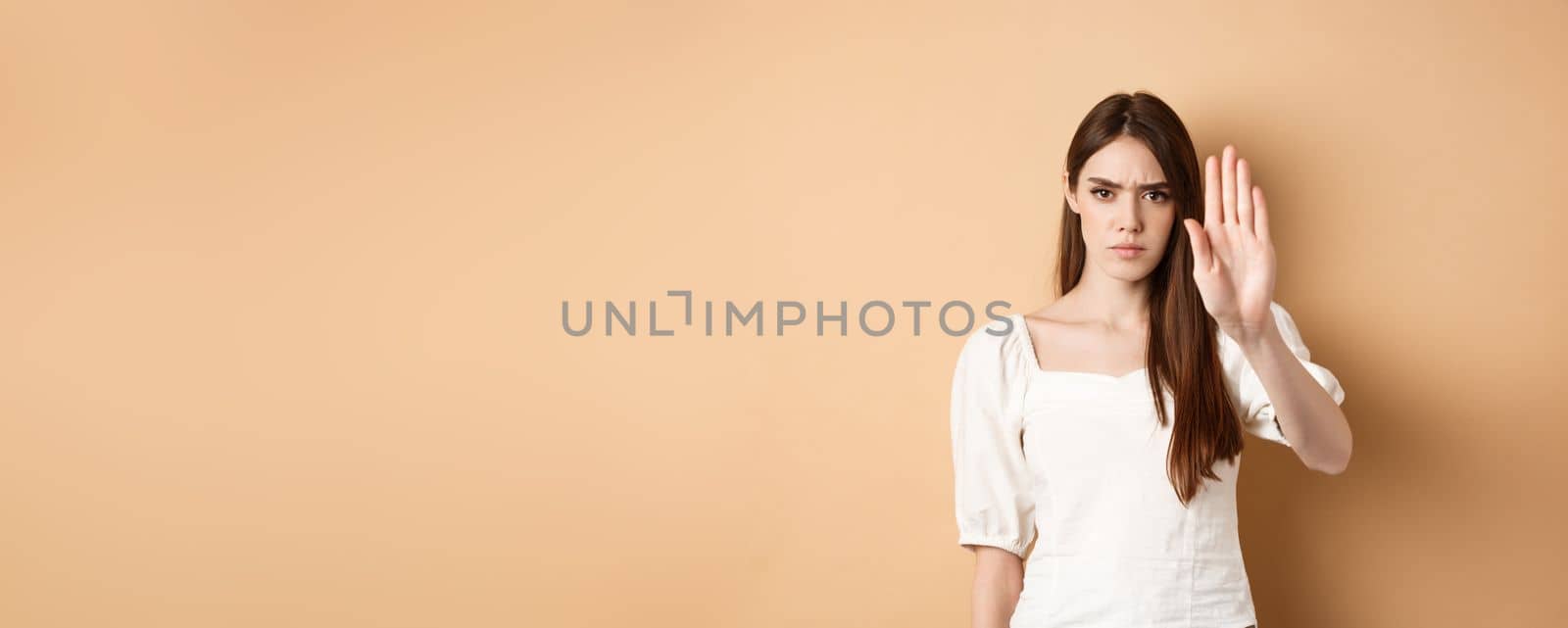 Nope stop right there. Serious and confident woman stretch out hand to forbid something, frowning and saying no, disagree and reject bad offer, standing on beige background by Benzoix