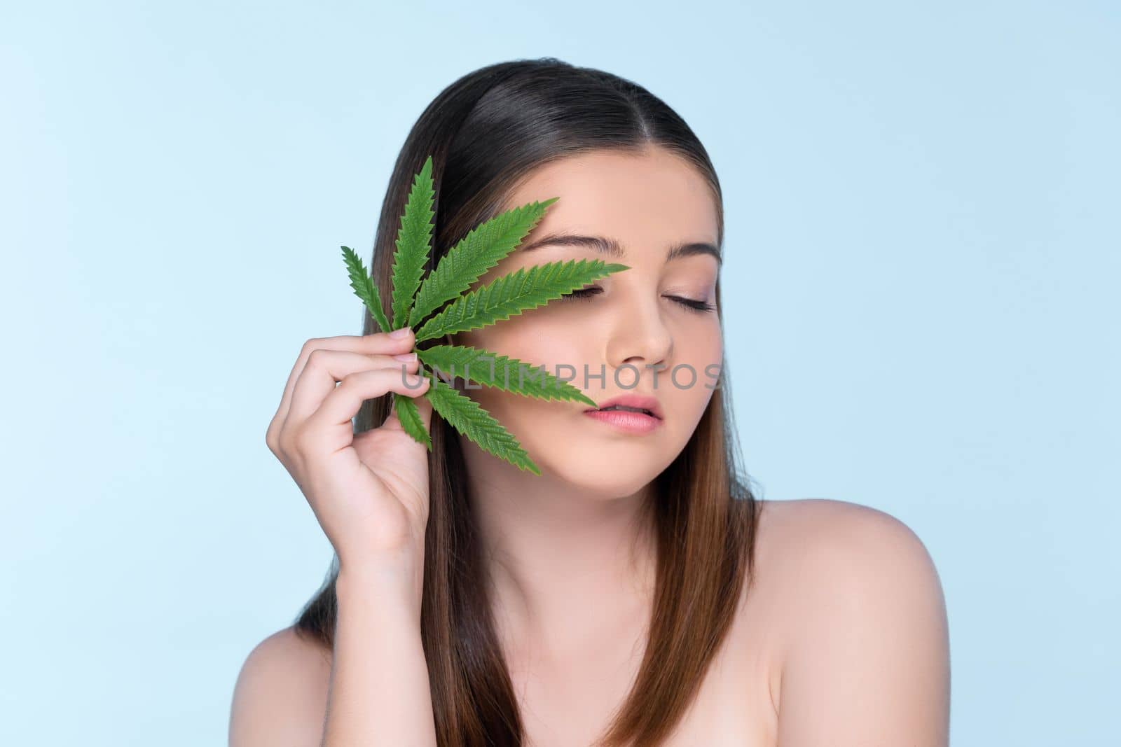 Closeup portrait of charming girl with fresh skin holding green leaf for beauty skin care made from cannabis leaf. Cosmetology and cannabis concept with isolated background.