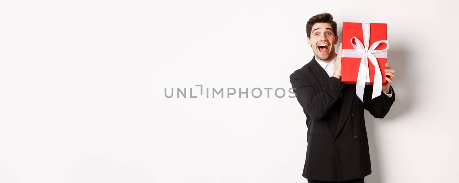 Image of happy good-looking man in black suit, holding box with christmas gift and smiling, standing against white background.
