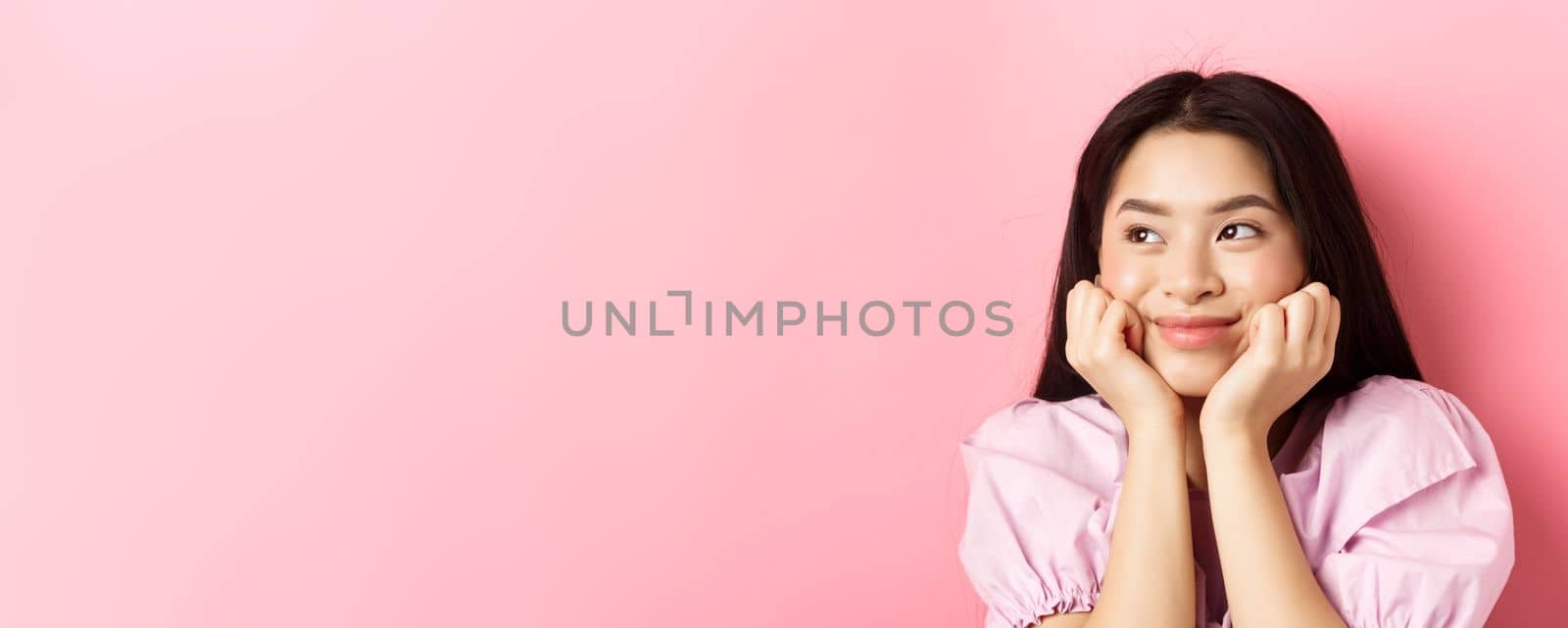 Close-up of excited asian girl listening with interest, smiling amused and looking aside at logo, daydreaming, standing against pink background by Benzoix