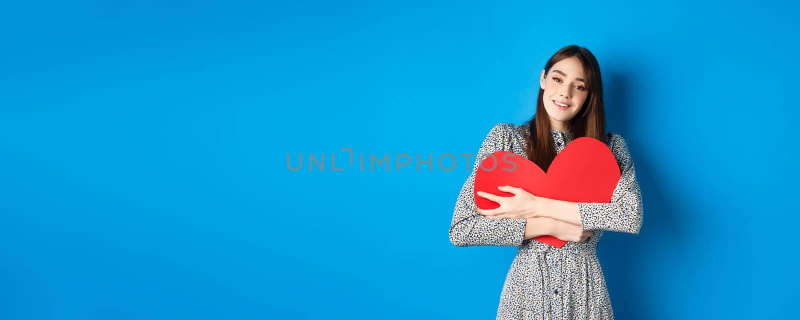 Valentines day. Dreamy romantic woman hugging big red heart cutout, looking sensual at camera, standing in dress on blue background.
