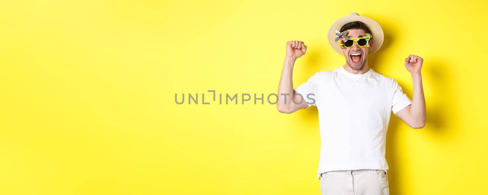 Concept of tourism and lifestyle. Happy man winning trip to resort, shouting yes and raising hands up, triumphing, wearing sunglasses and summer hat, yellow background.