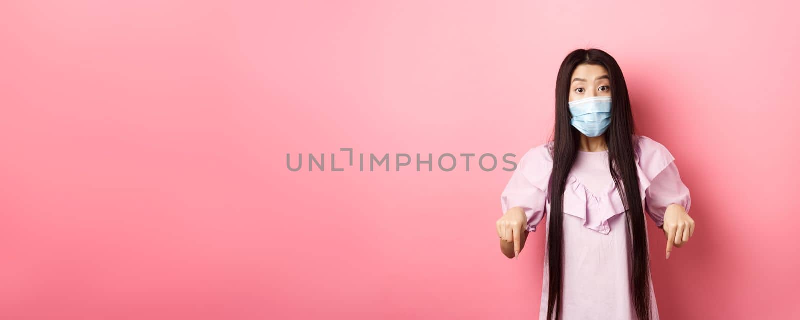 Healthy people and covid-19 pandemic concept. Excited asian girl in face mask pointing fingers down, showing advertisement with interest, standing against pink background by Benzoix