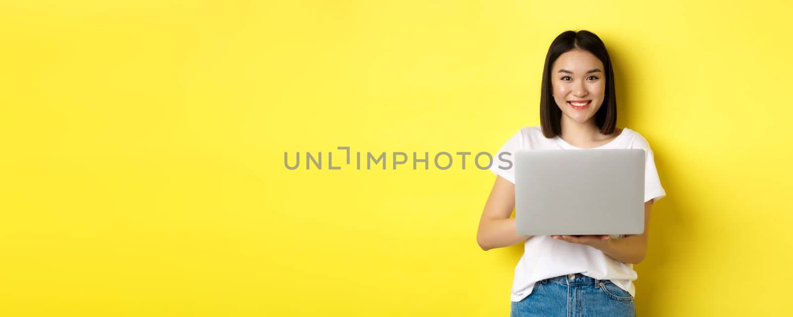 Cute asian woman studying on laptop and smiling, standing in white t-shirt and jeans against yellow background.