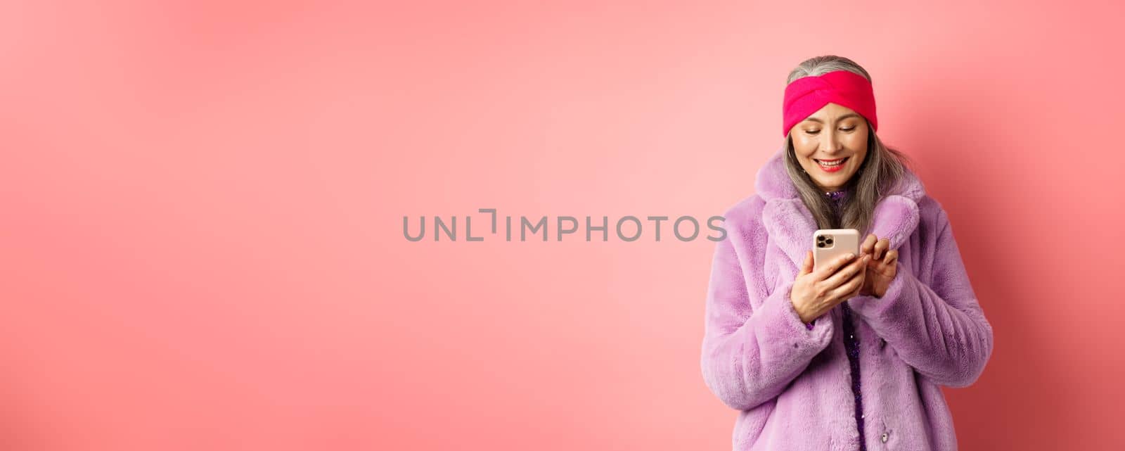 Online shopping and fashion concept. Modern asian granny in trendy faux fur coat texting message, using smartphone and looking happy at screen, pink background.