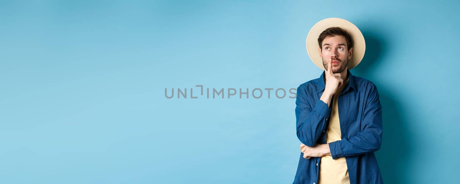 Thoughtful male tourist thinking of summer holiday vacation, looking aside with pensive face, touching lip and pondering next travel, standing on blue background.