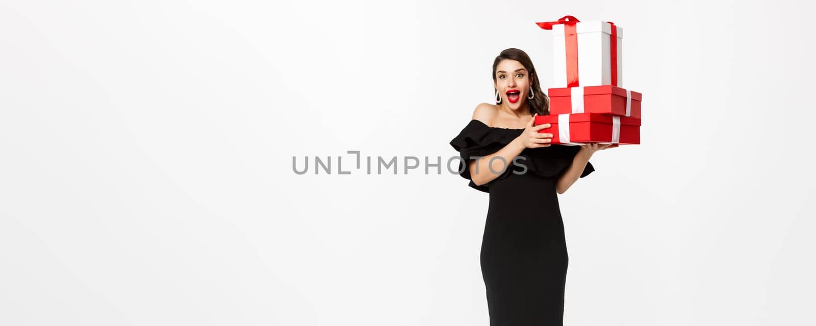 Merry christmas and new year holidays concept. Cheerful lady in black dress holding xmas presents and smiling at camera, standing over white background.