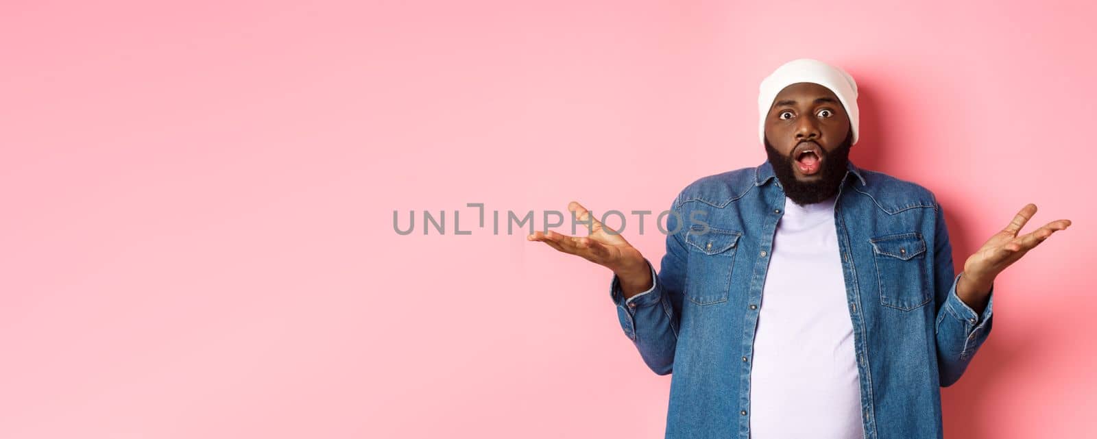 Confused and shocked african american man spread hands sideways and drop jaw, staring with awe and amazement at camera, standing over pink background clueless.
